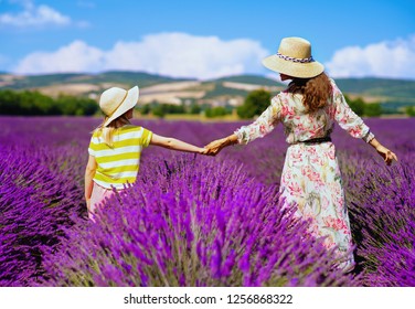 Seen From Behind Young Mother And Daughter In Lavender Field In Provence, France Stand Holding Hands. Family Having Agritourism Travel To The South Of France. Mother And Daughter Enjoying Summer