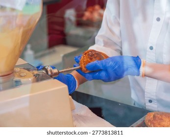 Seen through the subtle reflection of display glass, a bakery worker in blue gloves presents a golden apple turnover held in tongs, bridging the divide between kitchen and customer. The glass partition adds a voyeuristic layer to the service moment, framing the fresh pastry as both craft product and retail merchandise in a single composed image.