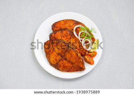 Seer fish fry arranged beautifully and garnished with onion, lemon and tomato slices on white ceramic plate with grey textured background.