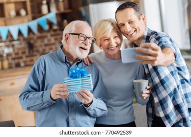 Seizing the moment. Cheerful young man taking selfies with his elderly parents during the celebration of the fathers birthday while the man holding a gift box