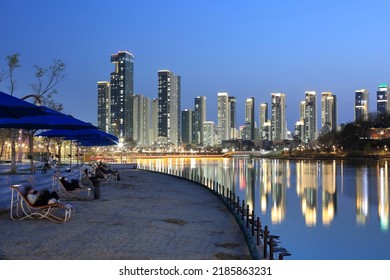 Sejong-si, Chungcheongnam-do, South Korea - April 9, 2022: Night View Of Sand And Tourists On Sunbed With Parasol Near Lake Against Apartments In The Background At Sejong Lake Park
