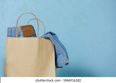 Select Focus Of Blue Jeans In A Paper Craft Bag On A Blue Background. Concept Of Thrift Stores, Resale, Second Hand. Copy Space.