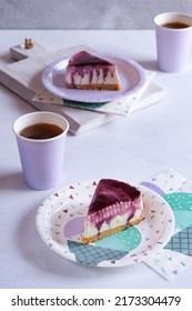 Selected Focus In Blueberry Cheesecake Slices On White Paper Party Plate With Blurred Purple Glass And Blurred Background Of Cheesecake