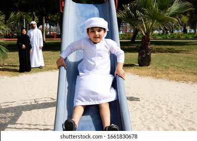 Selected Focus On Arab Child Boy. Arabic Kid Playing On Slide On A Park Wearing Kandura. Blurred Parents On The Background. Middle Eastern Locals