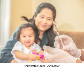 Selected Focus On A Doctor Hand With Plastic Gloves Holding A Hypodermic Syringe For Taking A Baby Vaccine, Mother And Baby As A Blurred Background