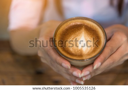 Selected focus on hot coffee cup. Women hands holding big  clear cup of cappuccino coffee blurred wooden table background. Women hold hot latte coffee cup &  art foam decorated on surface. Refreshment