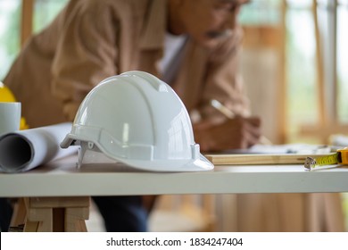 Selected Focus Of Safety White Hard Hat On Table Work Station And Blurred Background Of Senior Asian Man. Home Improvement DIY In Workshop.