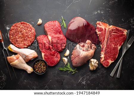 Selection of assorted raw meat food with seasonings for zero carb carnivore diet: uncooked beef steak, ground meat patty, heart, liver and chicken legs on black stone background from above 