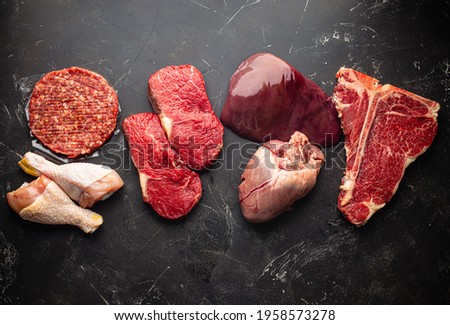 Selection of assorted raw meat food for zero carb carnivore diet: uncooked beef steak, ground meat patty, heart, liver and chicken legs on black stone background from above 