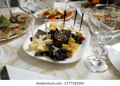 A selection of different cheeses is arranged on a white dish alongside dips and fruit. This food display is part of a larger spread at a gathering in the evening.