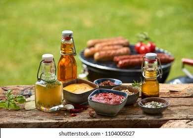 Selection of dressings, sauces, marinade and spices laid out on a rustic wooden garden table for a summer barbecue with sausages grilling over the fire in the background