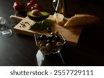 A selection of fresh olives and avocado slices on a cutting board with bread and tomatoes. Prepared for a healthy snack or appetizer on a dark, moody kitchen surface.