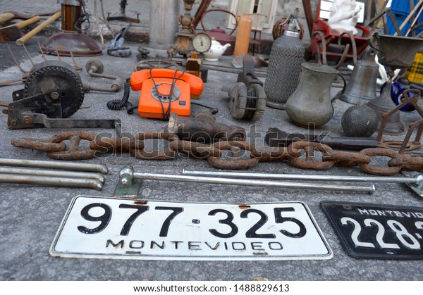 Selection of objects at a flea\
market laid out on the ground. Main features are a bright orange\
telephone, car license plate from Montevideo and a large rusted\
chain.