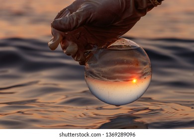 Selection Of Water Analysis In The Sea Or Lake. The Water Sample Contains Organic Garbage, Algae. A Gloved Hand Holds A Round Flask Of Water. The Sphere Shows The Horizon, Sunset And Blue Sky.