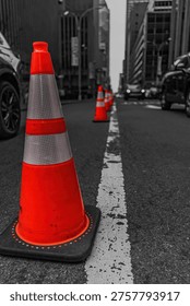 Selective color, vertical composition of vibrant orange traffic cones on a grayscale Midtown Manhattan street. A close-up of road safety equipment against a black-and-white urban background on an overcast day in New York City. Ideal for concepts of safety, urban development, caution, and city infrastructure.