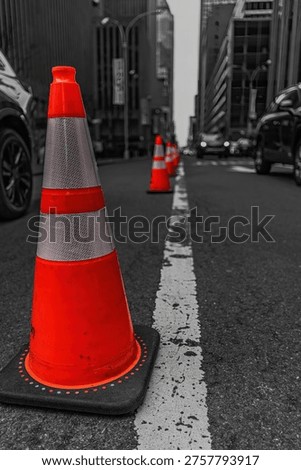 Selective color, vertical composition of vibrant orange traffic cones on a grayscale Midtown Manhattan street. A close-up of road safety equipment against a black-and-white urban background on an overcast day in New York City. Ideal for concepts of safety, urban development, caution, and city infrastructure.