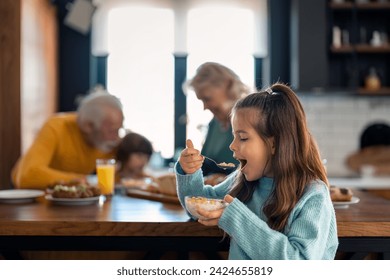 Selective focus of adorable small girl eating corn flakes cereals for breakfast while standing in the kitchen with grandparents and younger sibling in the background. - Powered by Shutterstock