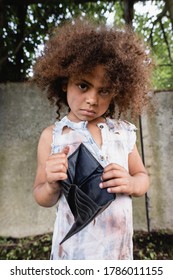 Selective Focus Of African American Child In Dirty Clothes Holding Empty Wallet And Looking At Camera On Urban Street