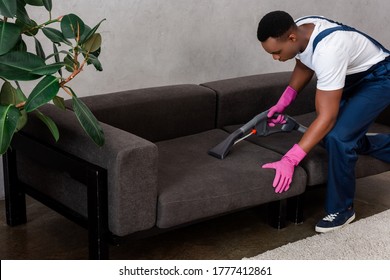 Selective Focus Of African American Cleaner In Uniform Cleaning Upholstery Of Couch At Home