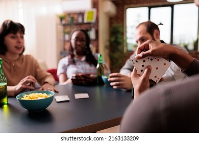 Selective focus of african american man holding cards while playing with best friends at home. Happy multiethnic people sitting at table in living room while enjoying fun leisure activity together. - Powered by Shutterstock