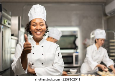 Selective focus of an African female baker in a chef uniforms, standing with arms folded while thumbs up and smiling at the camera, with blurred colleagues in the background. Copy space on right side. - Powered by Shutterstock