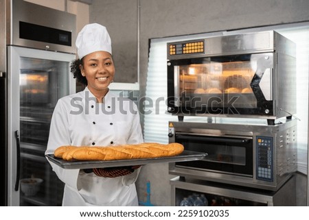 Selective focus of an African female baker in a chef uniform, standing holding a tray of freshly baked French breads and smiling at camera beside the oven that was baking breads in the bakery kitchen.