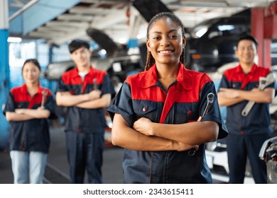 Selective focus of an African female mechanic in uniform, standing to hold a ring spanner with arms folded and smiling at camera with a blurred mechanic team standing in the background in the garage. - Powered by Shutterstock