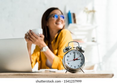 Selective Focus Of Alarm Clock And Happy Freelancer Having Coffee Break In Home Office With Laptop