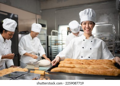 Selective focus of an Asian female baker in white chef uniform, standing holding a tray of freshly baked French breads and smiling at camera, with blurred colleagues kneading dough in the background.