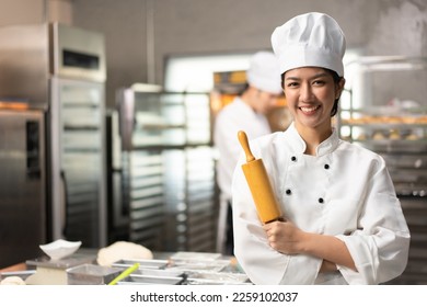 Selective focus of Asian female baker in white chef uniform, standing with arms crossed holding a rolling pin and smiling at camera, with blurred bakery kitchen in background. Copy space on left side. - Powered by Shutterstock