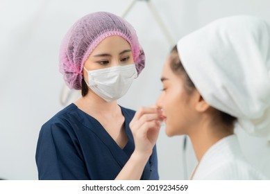 Selective Focus Of Asian Female Beautician In Blue Clothes, Medical Hat, And Face Mask Cleaning Facial Skin Of A Blurred Asian Female Client Before Doing A Beauty Procedure In Clinic Treatment Room. 