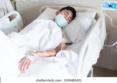 Selective Focus Asian Young Boy Sleeping On Hospital Bed During Treatment And Blurry Set Iv Fluid Intravenous Drop Saline.Young Man Is Sick With Protective Mask.Medical Treatment Concept.