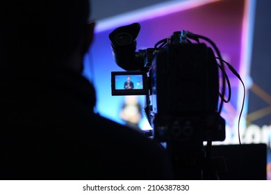 Selective focus of the back of the video camera capturing a blurred moderator image is on an opened LCD screen monitor while recording live at the event with the back of the cameraman in foreground.
