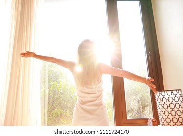 Selective Focus Of  Back View Young Woman In White Dress Which She Is Looking At Sunshine Day In The Morning And Stretch The Muscles At Window. Health Concept.