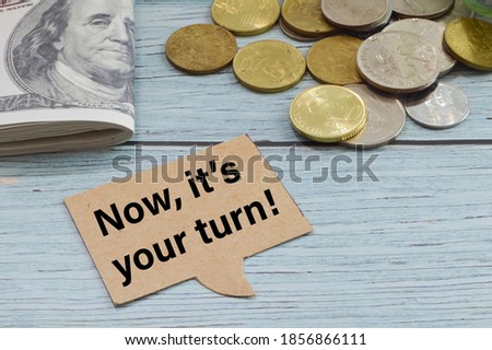 Selective focus of banknotes, stack of coins and memo note written with NOW IT'S YOUR TURN!. Business concept. 