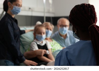 Selective Focus Of Black Asisstant Discussing With Caring Family Explaining Healthcare Treatment Against Clinical Disease In Hospital Ward. Elderly Senior Woman Resting In Bed Wearing Face Mask