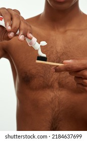 Selective Focus Of Black Guy Pouring Toothpaste On Toothbrush Before Brushing Teeth. Obscure Face Of Young Man With Naked Torso. Dental Care And Hygiene. Isolated On White Background. Studio Shoot