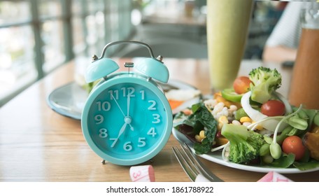 Selective focus of Blue clock with woman eating salad bowl with fruit after intermittent fasting sitting on a table at home - Shutterstock ID 1861788922