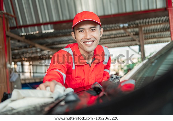 selective focus of car\
cleaner Asian male wearing red uniform smiling while wiping the car\
in the car salon
