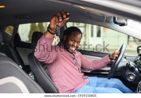 Selective focus at the car keys in hand of happy
African-American man. Cheerful black guy buying a car, got a
driving licence, sits and holds steering wheel and looks at the
camera, achieving
goal