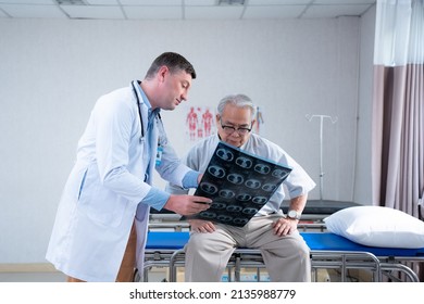 Selective Focus Of Caucasian Male Doctor In A White Medical Gown, Standing Pointed Into The Abdomen MRI Scan Film To Asian Senior Male Patient Sitting On Examination Bed In A Hospital Examination Room