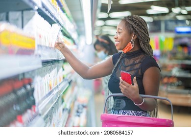 Selective Focus Of Cheerful African Lady, Carrying Basket In Her Arm- Black Woman In Front Of Shelf, Face Mask Lowered At Her Chin- Shopping Concept