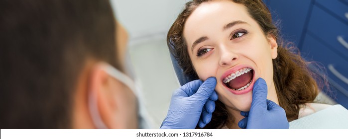 selective focus of cheerful woman in braces opening mouth during examination of teeth near dentist