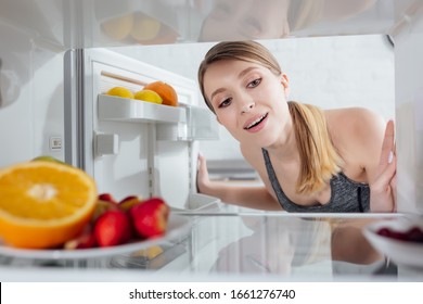 selective focus of cheerful woman looking at fruits in fridge