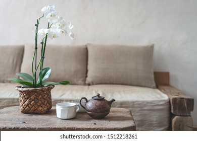 Selective focus of clay teapot, white cup and orchid flower at wooden table against blurred wall with copy space on background. Cozy house with comfort sofa in vintage interior style
