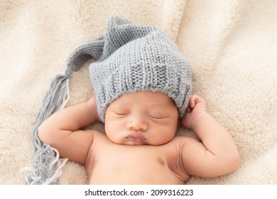 Selective Focus Close Up Of Asian Little Baby Sleeping On Back With His Hands Behind His Head On Beige Blanket, Adorable Toddler Wearing Knitted Gray Hat. Infant Sleeping On Bed With Happy And Safe.