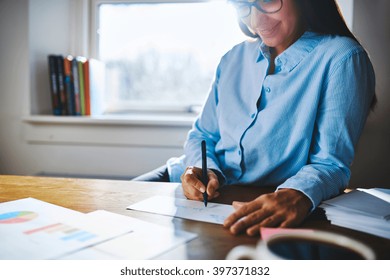 Selective Focus Close Up Of Cheerful Young Self-employed Woman Wearing Glasses And Blue Shirt At Desk Writing On Form Next To Cup Of Coffee And Booklets