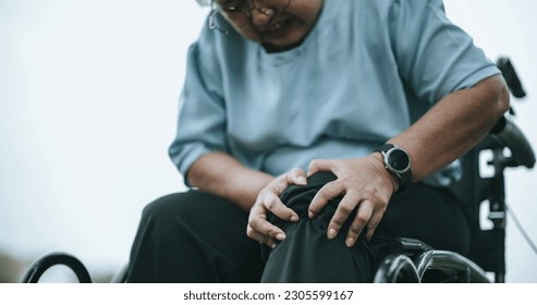 Selective focus, Close up, Asian Mature woman in wheelchair having knee with pain expression on face, She Use both hands to press on the painful area. healthcare in retirement age concept