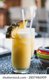 A selective focus close-up shot of two tropical yellow cocktails served in tall glasses with drinking straws. Each drink is topped with a thick white foam layer and garnished with a slice of fresh pineapple, placed on a dark tray in an outdoor terrace restaurant.