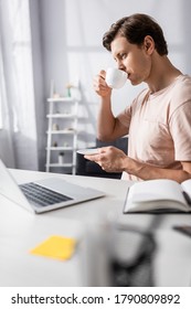 Selective focus of concentrated freelancer looking at laptop while drinking coffee at home, earning online concept